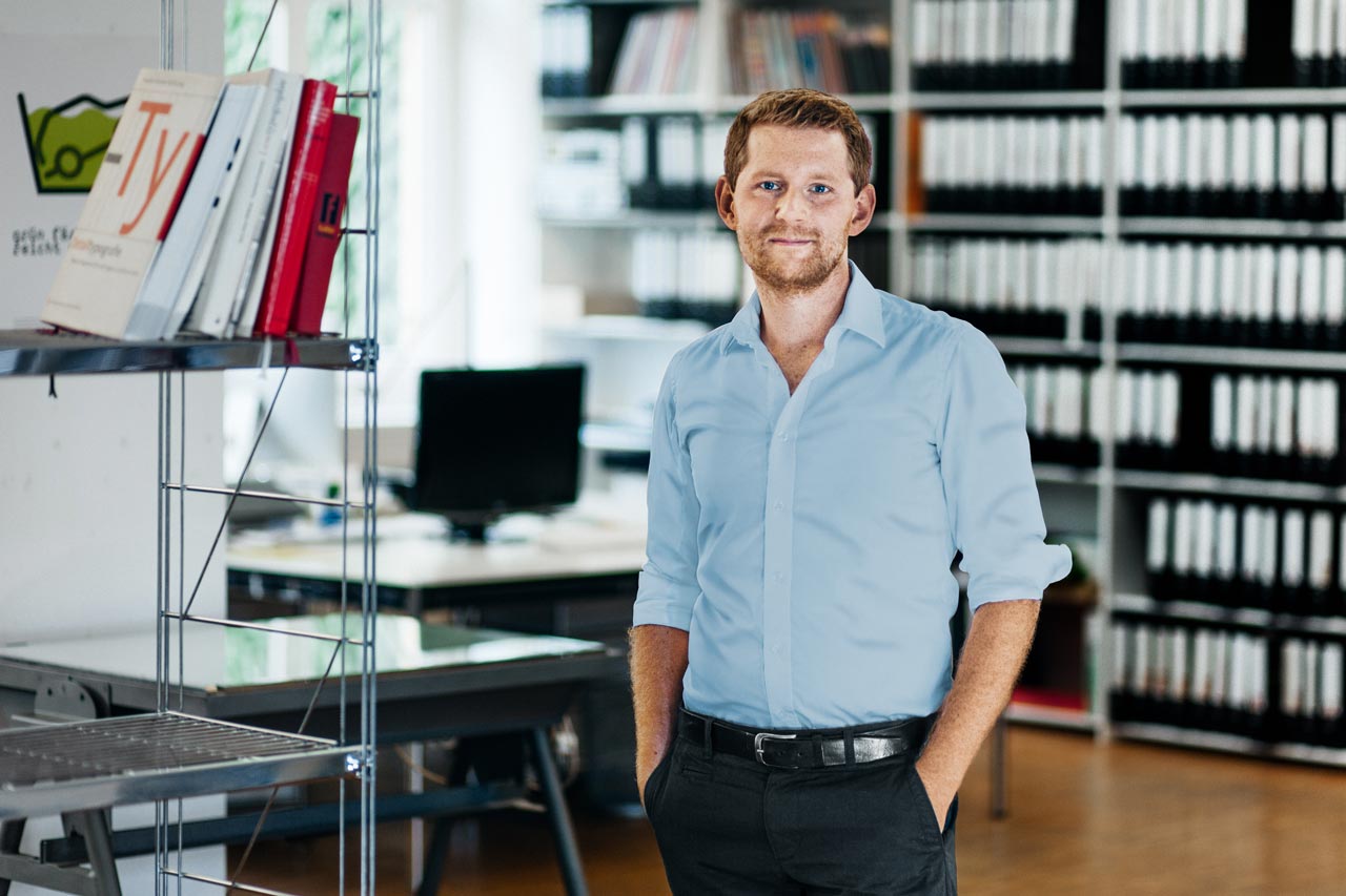 Gleicher Bildinhalt wie im ersten Bild: Ein junger Mann steht in einem Büro. Im Hintergrund steht ein Schreibtisch mit einem Monitor und Regal mit vielen Aktenordnern.