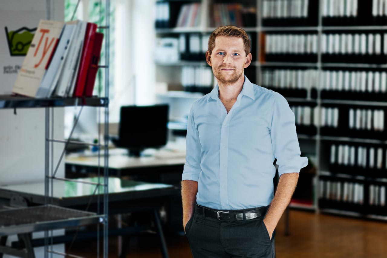 Gleicher Bildinhalt wie im ersten Bild: Ein junger Mann steht in einem Büro. Im Hintergrund steht ein Schreibtisch mit einem Monitor und Regal mit vielen Aktenordnern.